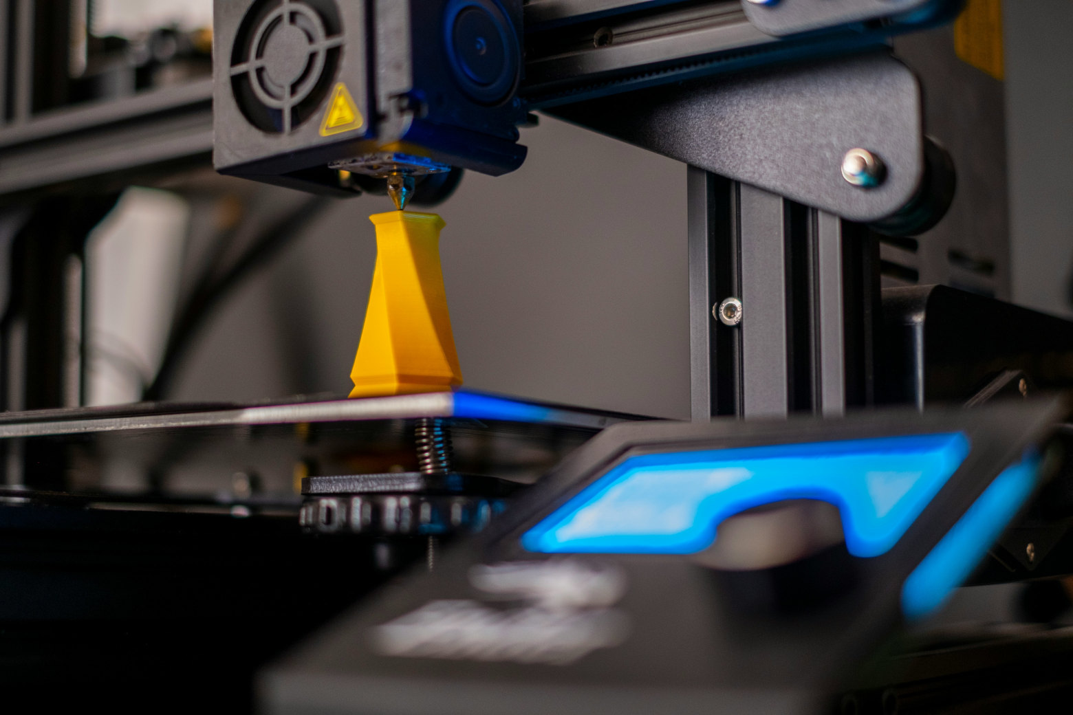 Dark 3D-printer with glowing blue controller printing yellow tower. Courtesy of Osman Talha Dikyar and used under the unsplash license.